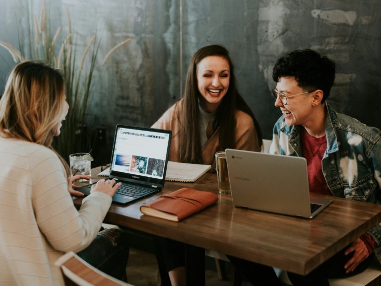 Three people are engaged in a lively discussion at a table with laptops and drinks.