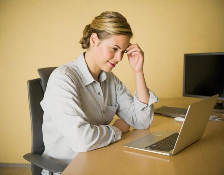 Woman looking pensive while working on a laptop at a desk in an office setting.